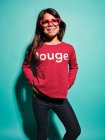 Happy preteen girl in pink heart shape glasses and casual clothes smiling away while standing with hands on hips against turquoise background in modern studio — Stock Photo