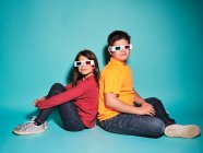 Side view of cheerful preteen boy and girl in casual clothes and three dimensional glasses looking at camera sitting back to back against blue background — Stock Photo