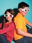Вид збоку веселий хлопчик і дівчинка в повсякденному одязі і тривимірні окуляри, що сидять на спині на синьому фоні — стокове фото