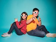 Side view of cheerful preteen boy and girl in casual clothes and three dimensional glasses pointing and looking at camera sitting back to back against blue background — Stock Photo