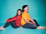 Side view of cheerful preteen boy and girl in casual clothes and three dimensional glasses sitting back to back against blue background — Stock Photo
