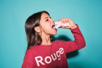 Side view of happy little girl in red casual wear eating pink soft candy marshmallow looking away while standing against blue background — Stock Photo