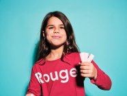 Happy little girl in red casual wear eating pink soft candy marshmallow looking at camera while standing against blue background — Stock Photo
