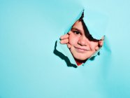 Cheerful excited little boy with grimace looking at camera through ripped colorful blue paper sheet in studio — Stock Photo