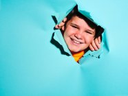 Joyeux petit garçon excité avec grimace regardant la caméra à travers déchiré feuille de papier bleu coloré en studio — Photo de stock