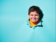 Cheerful excited little boy with grimace looking at camera through ripped colorful blue paper sheet in studio — Stock Photo