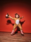 Energique petite fille heureuse en costume de cerf mignon écartant les bras tout en sautant la recherche contre le mur rouge en studio — Photo de stock
