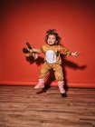Enérgica niña feliz en traje de ciervo lindo brazos extendidos mientras salta mirando hacia arriba contra la pared roja en el estudio - foto de stock