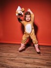 Енергійний щасливий маленька дівчинка в милий костюм оленя, розтягуючи руки, стрибаючи, дивлячись на червону стіну в студії — стокове фото