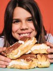 Closeup portrait of cheerful little girl in casual wear enjoying sweet eclairs with chocolate while sitting at table against red background — Stock Photo
