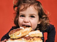 Nahaufnahme Porträt eines fröhlichen kleinen Mädchens, das süße Eclairs mit Schokolade genießt, während es vor rotem Hintergrund in die Kamera blickt — Stockfoto