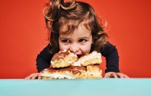 Nahaufnahme Porträt eines fröhlichen kleinen Mädchens, das süße Eclairs mit Schokolade genießt, während es auf dem Tisch vor rotem Hintergrund sitzt und wegschaut — Stockfoto