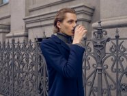Серйозний молодий чоловік зі стильним зачіскою в модному пальто виймає каву, стоячи біля старого металевого паркану проти кам'яної будівлі в місті — стокове фото