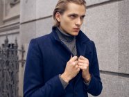 Вид сбоку стильный молодой мужчина с модной прической, одетый в элегантное пальто, стоящее против серой каменной стены в городе — стоковое фото