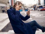 Side view of young handsome male in stylish outfit looking at smartphone and brushing hair while sitting on city street in gray autumn day — Stock Photo