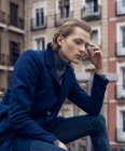 Low angle side view of serious elegant young male in stylish coat leaning on hand and thinking while sitting on street against blurred building — Stock Photo