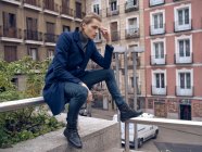 Low angle side view of serious elegant young male in stylish coat leaning on hand and thinking while sitting on street against blurred building — Stock Photo