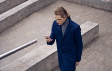 Dall'alto di serio giovane uomo d'affari in elegante cappotto controllo messaggio sul telefono cellulare mentre in piedi su scala in pietra sulla strada della città — Foto stock