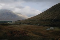 Шотландский пейзаж с узкой извилистой рекой, протекающей среди покрытых травой холмов под облачным небом в осенний день — стоковое фото