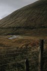 Schottische Landschaft mit Drahtzaun mit schmalem, geschwungenem Fluss, der zwischen mit Gras bedeckten Hügeln unter wolkenverhangenem Himmel im Herbst fließt — Stockfoto