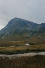 Tranquil scenery of Scottish countryside with yellow grassland and lonely small house located near rocky mountain and small stream river against cloudy sky — Stock Photo