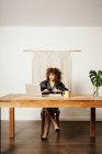 Eine erwachsene Dame in Lederjacke und mit lockigem Haar sitzt am Tisch gegen Makrammedekoration und liest während der Projektarbeit am Arbeitsplatz Daten auf dem Laptop — Stockfoto