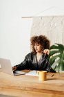 Взрослая женщина в кожаной куртке и с вьющимися волосами сидит за столом против макраме украшения и чтения данных на ноутбуке во время работы над проектом на рабочем месте — стоковое фото