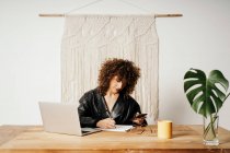 Femme d'affaires rétro positive aux cheveux bouclés souriant et écrivant dans un cahier assis à table et utilisant un smartphone au bureau — Photo de stock