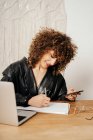Позитивна ретро-бізнес-леді з кучерявим волоссям, посміхаючись і пишучи в блокноті, сидячи за столом і використовуючи смартфон в офісі — стокове фото