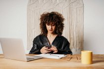 Positive retro businesswoman with curly hair sitting at table and using smartphone in office — Stock Photo