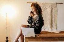 Вид на счастливую женщину-менеджера в ретро-костюме, сидящую на столе рядом с ноутбуком и отвечающую на телефонный звонок во время работы в освещенном рабочем пространстве — стоковое фото