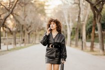 Emprendedora de cuerpo completo en traje vintage caminando por el camino de asfalto y teniendo una conversación de teléfonos inteligentes mientras viaja al trabajo a través del parque de la ciudad - foto de stock