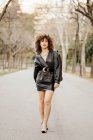 Empresária otimista em jaqueta de couro da moda e com cabelo encaracolado sorrindo para a câmera enquanto está em pé no fundo borrado da rua — Fotografia de Stock