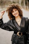 Optimistic female entrepreneur in trendy leather jacket and with curly hair smiling at camera while standing on blurred background of street — Stock Photo
