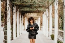 Junge Geschäftsfrau in retro-schwarzer Lederjacke und Rock surft im Smartphone, während sie vor der Arbeit in einer Bogenpassage im Park steht — Stockfoto