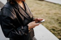 Junge Geschäftsfrau in retro-schwarzer Lederjacke und Rock surft im Smartphone, während sie vor der Arbeit in einer Bogenpassage im Park steht — Stockfoto