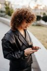 Young businesswoman in retro black leather jacket and skirt browsing smartphone while standing in arched passage in park before work — Stock Photo