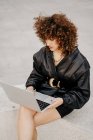 Сверху деловая женщина в черном кожаном костюме сидит на границе и просматривает данные на ноутбуке во время работы над удаленным проектом на городской улице — стоковое фото