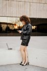 Side view of smart lady in vintage outfit browsing smartphone and standing near border with laptop while doing remote job on street — Stock Photo