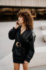 Все тело женщины-предпринимателя в винтажном наряде прогуливаясь по асфальтовой дорожке и разговаривая по смартфону во время поездки на работу через городской парк — стоковое фото