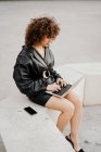 Intelligente donna d'affari riccia indossa vestito di pelle nera e giacca digitando sulla tastiera del computer portatile mentre seduto sulle scale e lavorando su un progetto remoto sulla strada della città — Foto stock