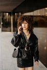 Умная деловая женщина в черном кожаном костюме и с кудрявыми волосами, отворачивающаяся и отвечающая на телефонный звонок, стоя рядом со зданием со стеклянной стеной на городской улице — стоковое фото