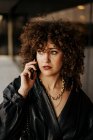Smart businesswoman in black leather suit and with curly hair looking away and answering phone call while standing near building with glass wall on city street — Stock Photo