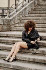 Полноразмерная стройная предпринимательница в кожаной юбке и куртке сидит на бетонных ступеньках и просматривает смартфон на городской улице — стоковое фото