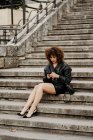 Empreendedor feminino magro de comprimento total em saia de couro e jaqueta sentado em passos concretos e smartphone de navegação na rua da cidade — Fotografia de Stock