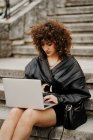 Inteligente mujer de negocios rizado con traje de cuero negro y chaqueta escribiendo en el teclado del ordenador portátil mientras está sentado en las escaleras y trabajando en el proyecto remoto en la calle de la ciudad - foto de stock