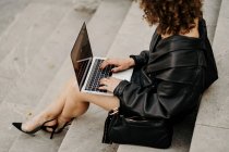 Empresária encaracolada inteligente vestindo terno de couro preto e jaqueta digitando no teclado do laptop enquanto se senta nas escadas e trabalha em projeto remoto na rua da cidade — Fotografia de Stock
