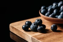 Frische reife Blaubeeren auf Holztisch neben Schüssel mit Beeren vor schwarzem Hintergrund platziert — Stockfoto