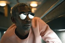 Взрослый афроамериканец в розовой толстовке и стильных солнцезащитных очках смотрит в сторону, опираясь на стену в освещенном туннеле — стоковое фото