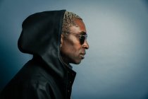 Side view of adult African American male in hooded coat and trendy sunglasses standing against blue background — Stock Photo